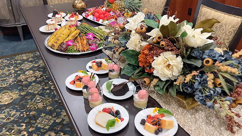 A dining table set with various plated desserts including slices of cake, fruit garnishes, and small glasses of mousse or pudding, alongside a large decorative floral arrangement with white, orange, and blue flowers and greenery.