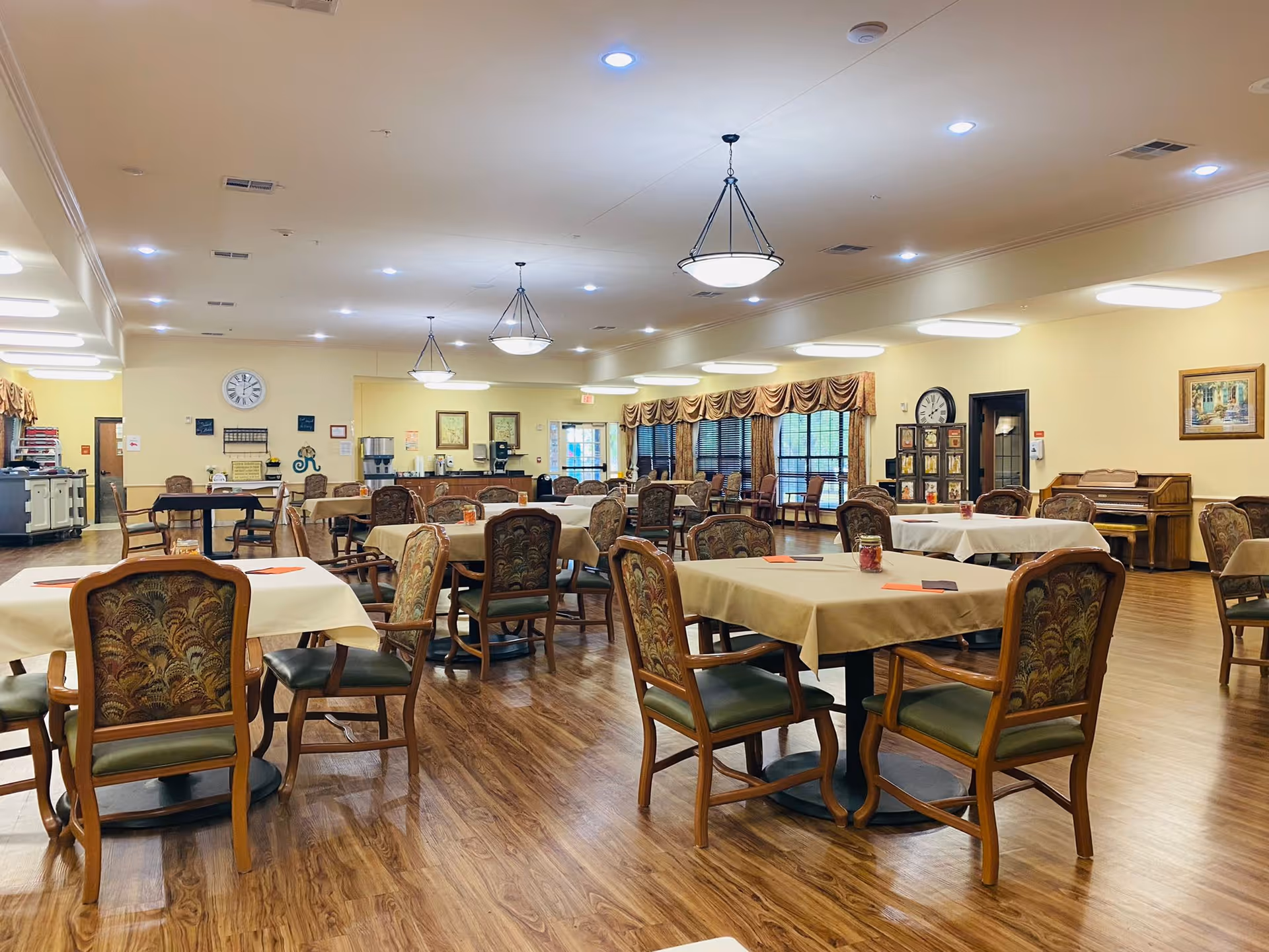 Spacious dining area with multiple tables covered in beige cloths, upholstered wooden chairs, pendant lights, and hardwood floors.
