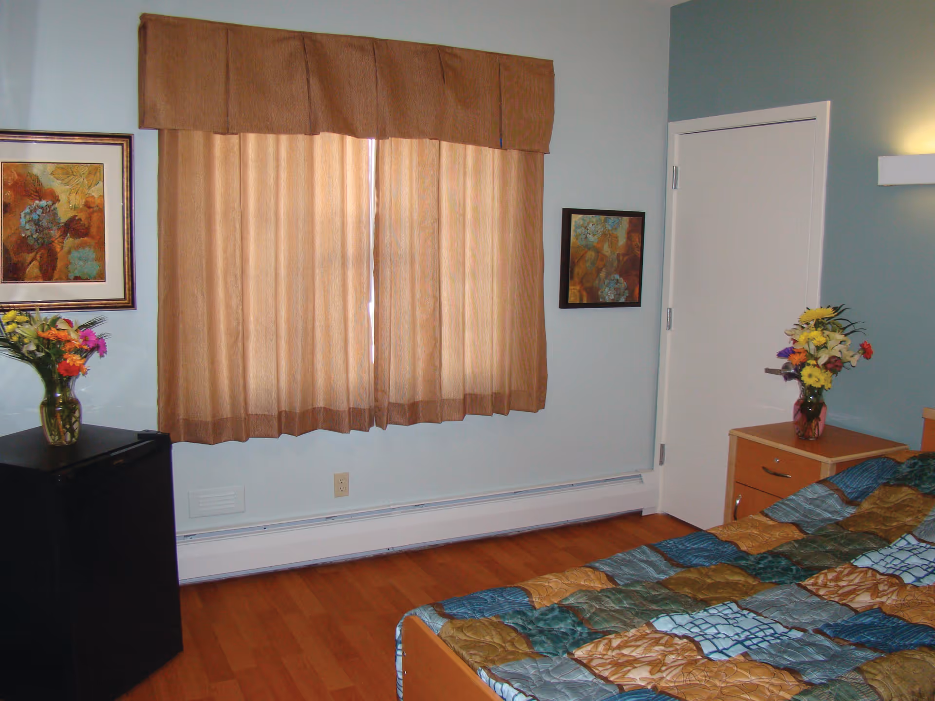 A bedroom with a bed covered in a multicolored quilt, a wooden nightstand with a vase of flowers, a window with beige curtains, two framed floral paintings on the walls, and a small black refrigerator with another vase of flowers on top.