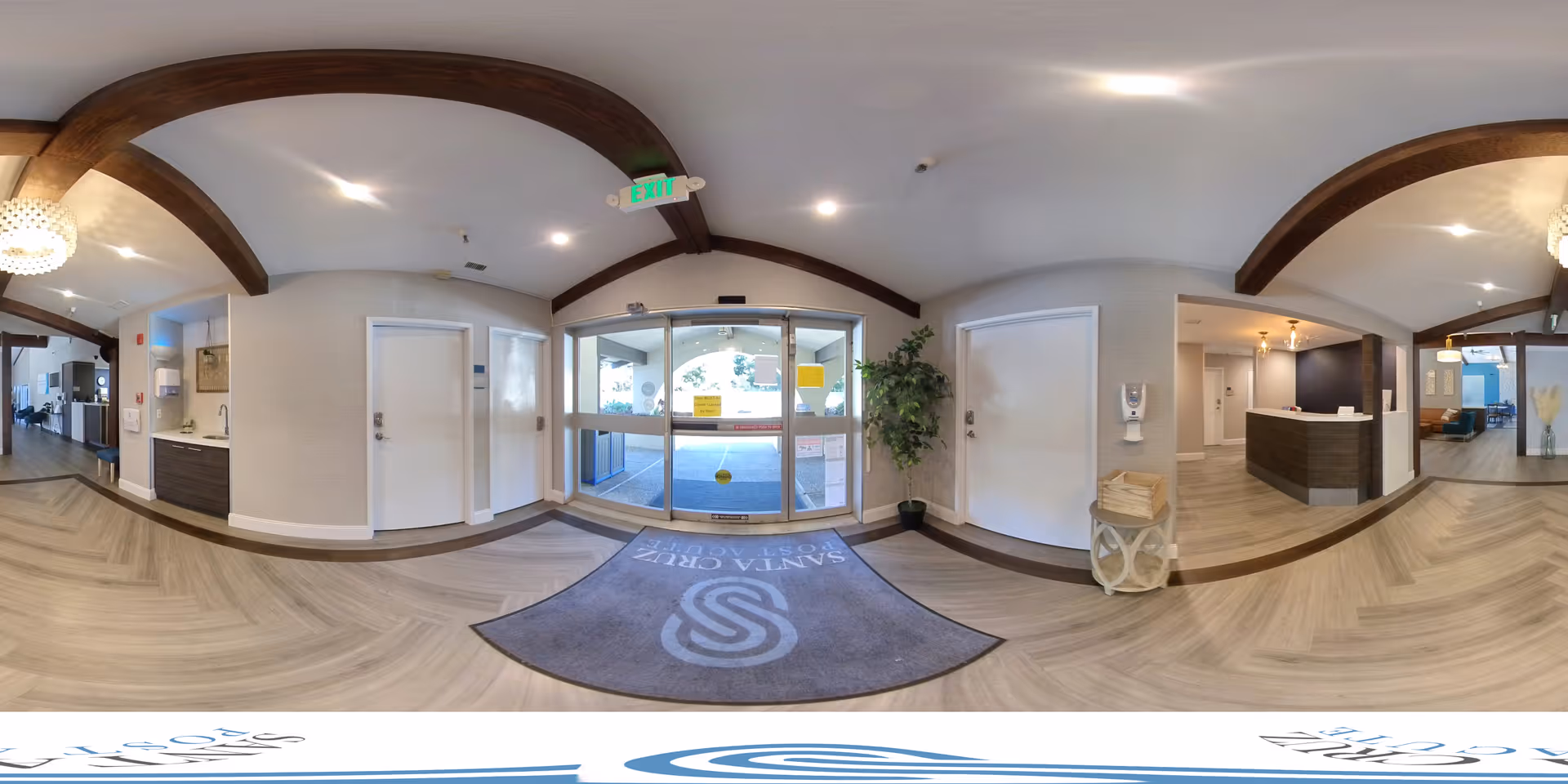 Panoramic view of the entrance lobby of Santa Cruz Post Acute facility featuring automatic sliding glass doors, a welcome mat with the facility logo, a reception desk to the right, a small sink area to the left, wood beam ceiling accents, and light wood flooring.