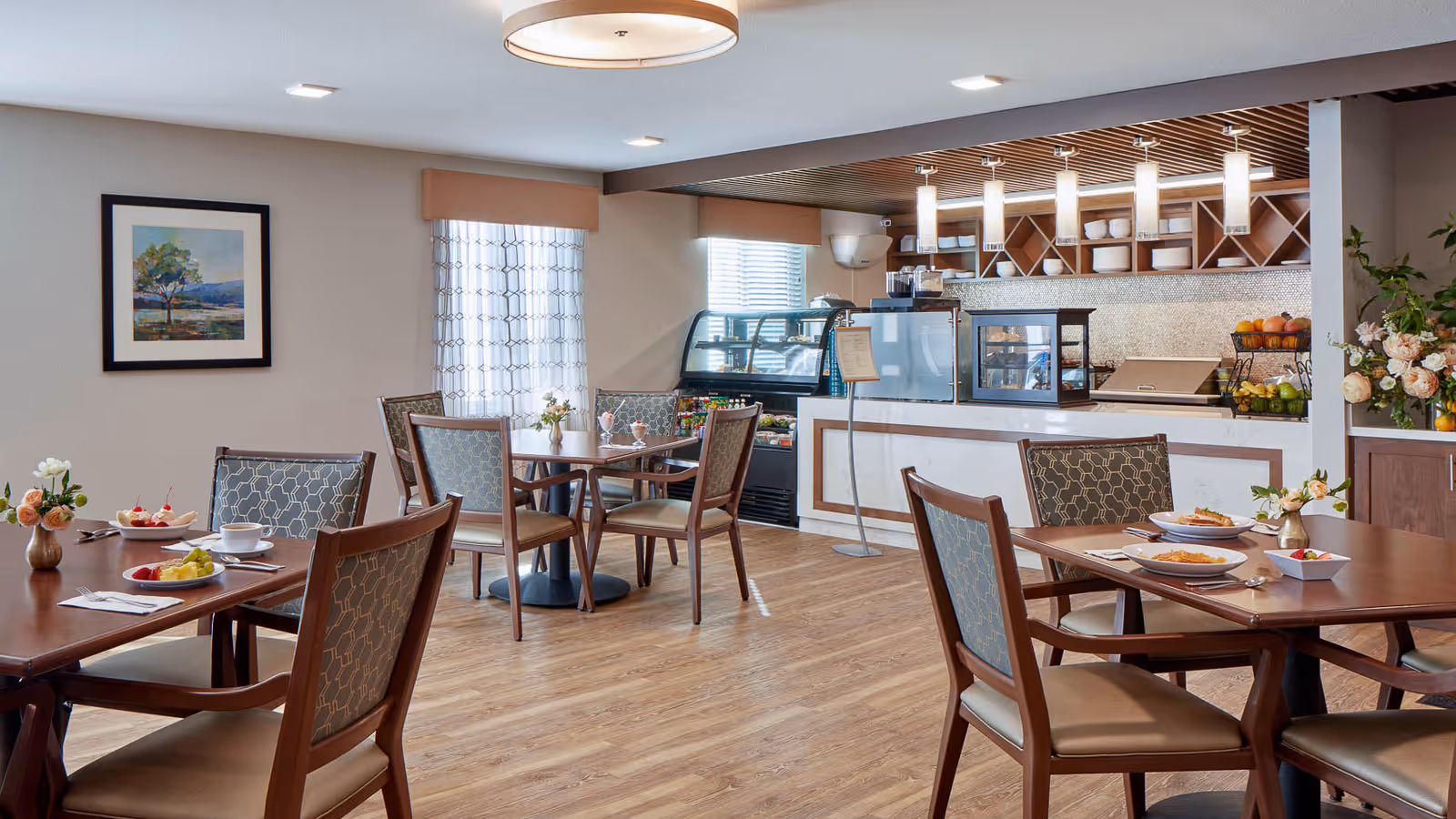 A dining area in a senior living facility with several wooden tables and chairs. The tables are set with plates of food, cups, and small flower arrangements. In the background, there is a counter with display cases containing food items and shelves with dishes. The room has wooden flooring, a framed painting on the wall, and a window with patterned curtains.