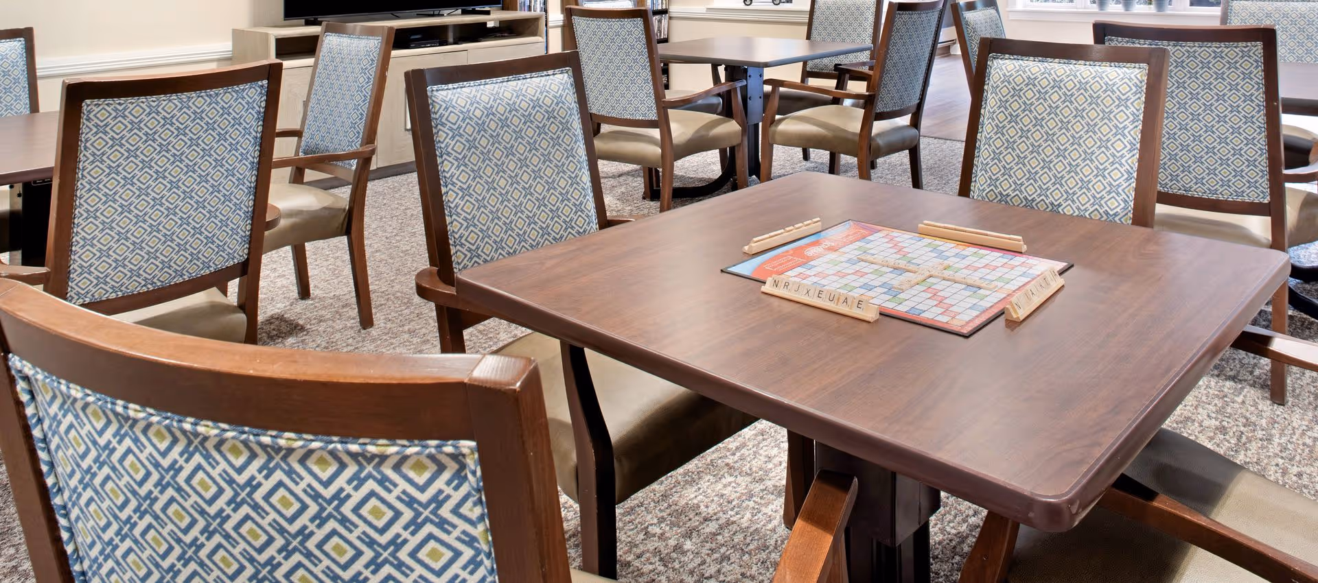 A room with several wooden tables and chairs with patterned upholstery. One table has a Scrabble board game set up on it. The room has carpeted flooring and light-colored walls with windows in the background.