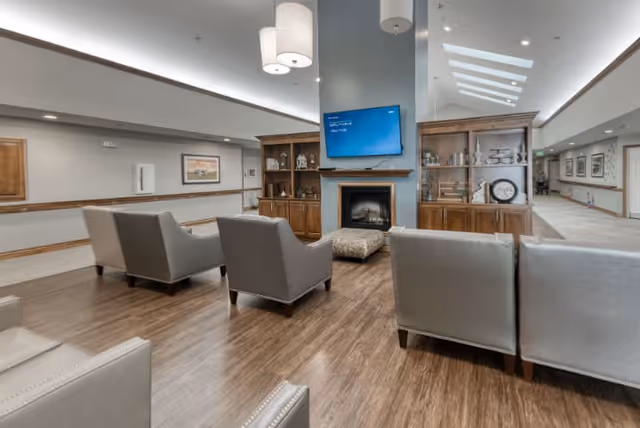 A spacious senior living facility common area with several gray armchairs arranged around a central ottoman. There is a fireplace with a mounted flat-screen TV above it, flanked by wooden built-in shelves displaying decorative items. The room has wood flooring, pendant lights hanging from the ceiling, and a hallway leading to other parts of the facility.