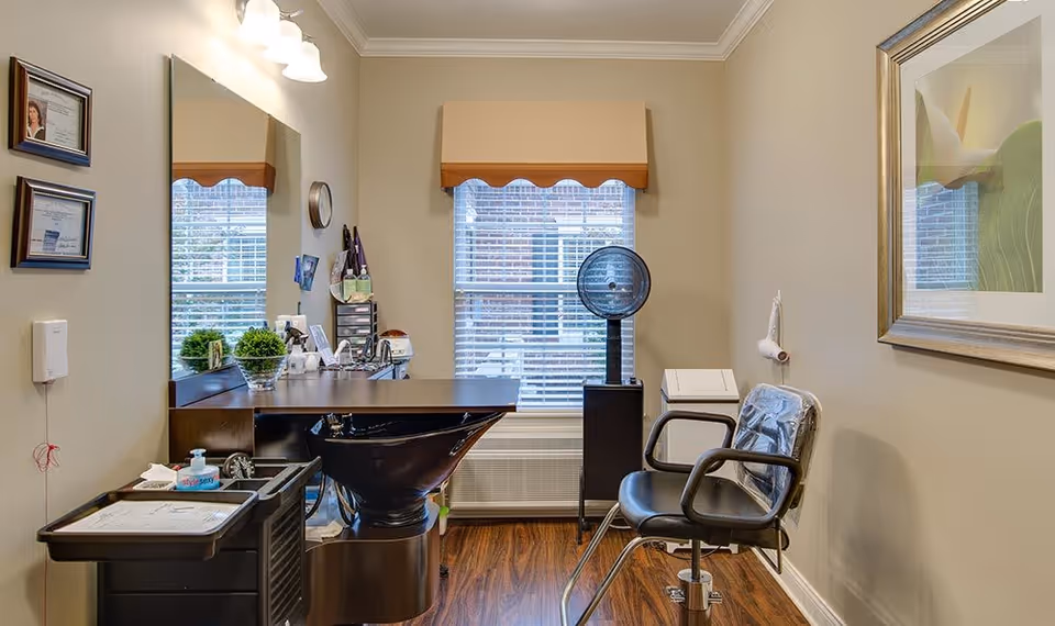 Small salon-style room with a styling station, shampoo bowl, salon chairs, mirror and a window with blinds.