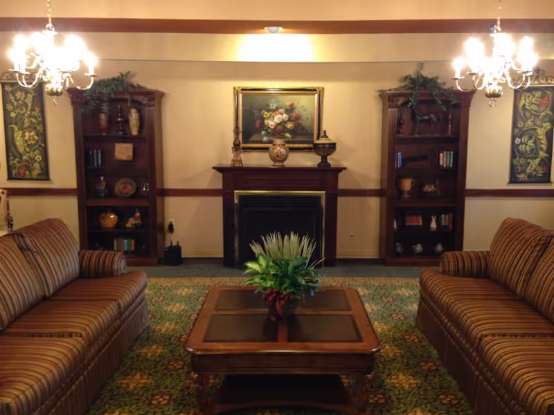 A cozy living room with two striped sofas facing each other across a wooden coffee table with a plant centerpiece. The room features a fireplace with decorative vases and a floral painting above it. On either side of the fireplace are wooden bookshelves filled with books and decorative items. Two chandeliers hang from the ceiling, and the floor is covered with a patterned carpet.