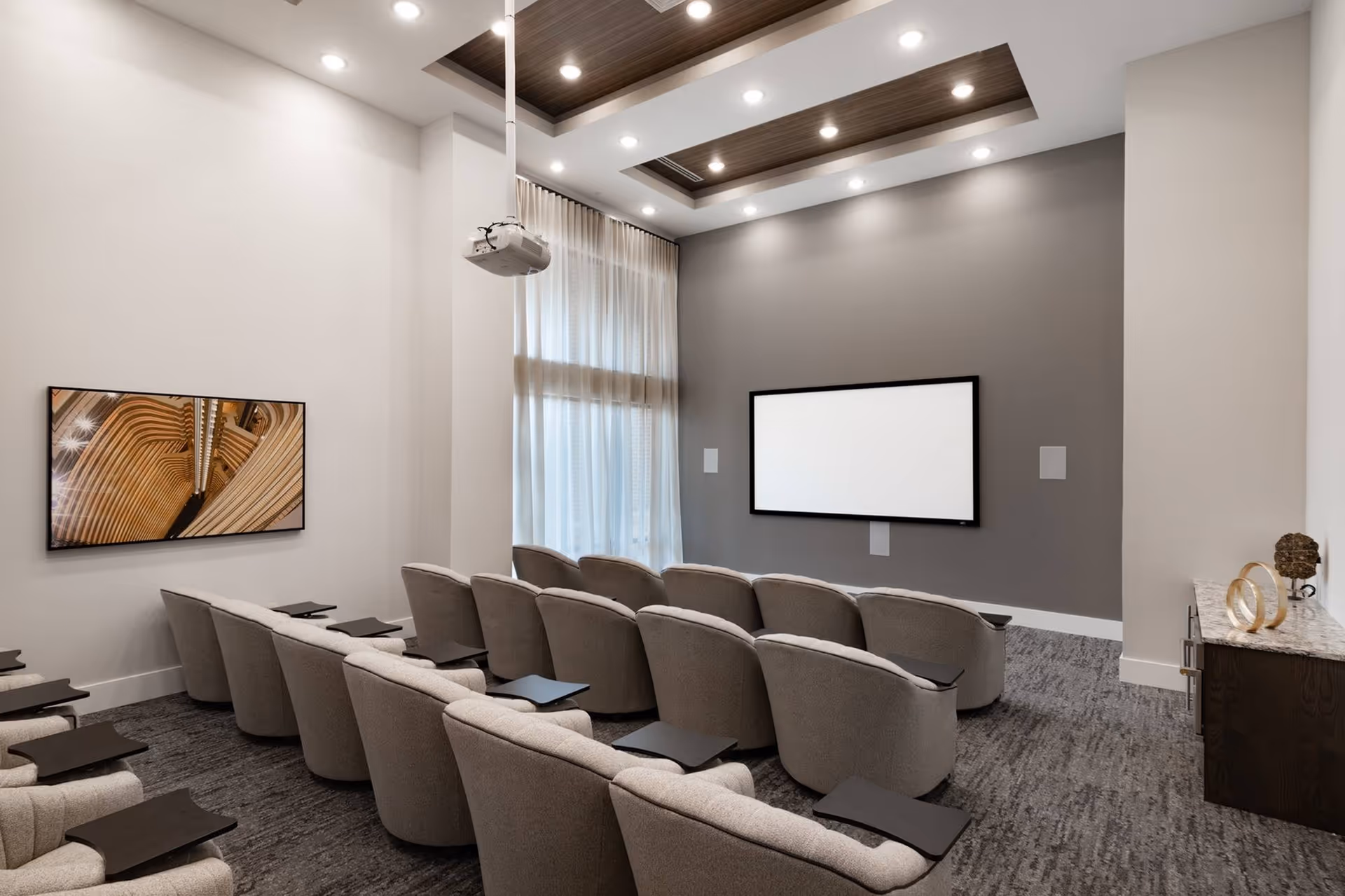 A modern media room with three rows of comfortable gray upholstered chairs, each equipped with a small black tray table. The room features a large blank projection screen on a gray accent wall, a ceiling-mounted projector, and a wall-mounted flat screen displaying an abstract image. The ceiling has recessed lighting and wooden panel accents, and sheer curtains cover a tall window on one side.