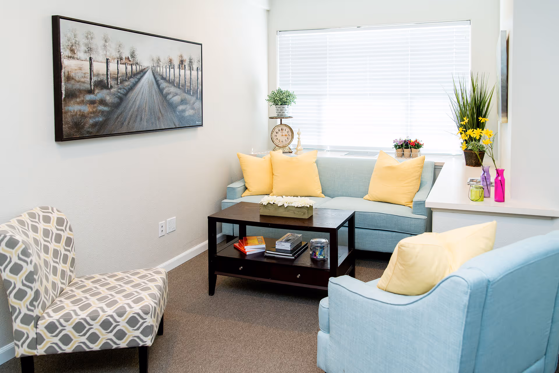 A bright, cozy living room with light-blue sofas and chair, yellow throw pillows, a dark coffee table, wall art, and a window with potted plants.