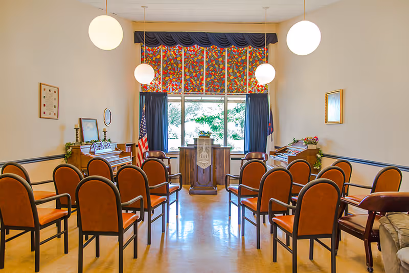 A small chapel or meeting room with rows of brown chairs arranged facing a wooden podium. There are two organs on either side of the podium, an American flag and a Christian flag near the window, and stained glass window panels above the large window letting in natural light. The room has white walls, hanging globe lights, and framed pictures on the walls.