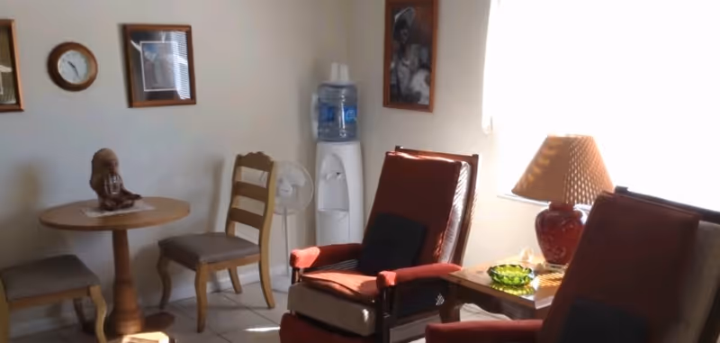 A cozy interior room with two cushioned armchairs, a small round wooden table with two chairs, a water dispenser, a standing fan, a table lamp on a side table, and framed pictures on the wall.