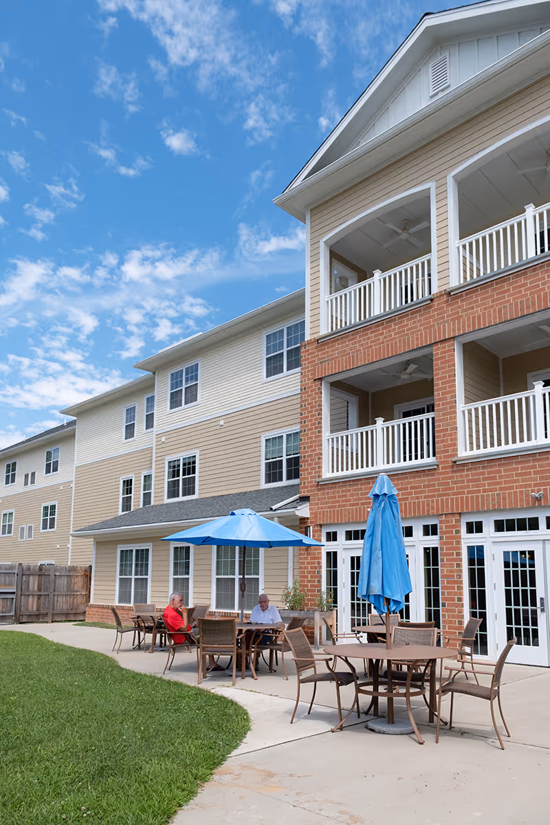 Outdoor patio area at a senior living facility with several tables and chairs, some shaded by blue umbrellas. Two elderly men are seated at one of the tables. The building has beige siding with red brick accents and white railings on balconies above. The sky is partly cloudy and blue.
