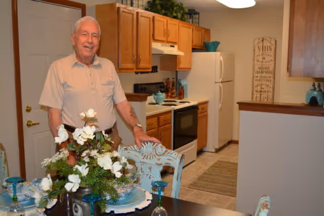 An elderly man standing in a kitchen area of a senior living facility named Spring Mill Meadows. The kitchen features wooden cabinets, a white refrigerator, a stove, and a decorative sign on the wall. In the foreground, there is a dining table set with plates, glasses, and a floral centerpiece.
