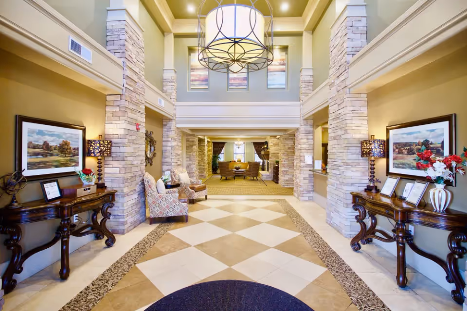 Bright and spacious senior living facility lobby with high ceilings, stone pillars, patterned tile floor, two ornate wooden tables with lamps and framed artwork on either side, and comfortable seating area in the background.