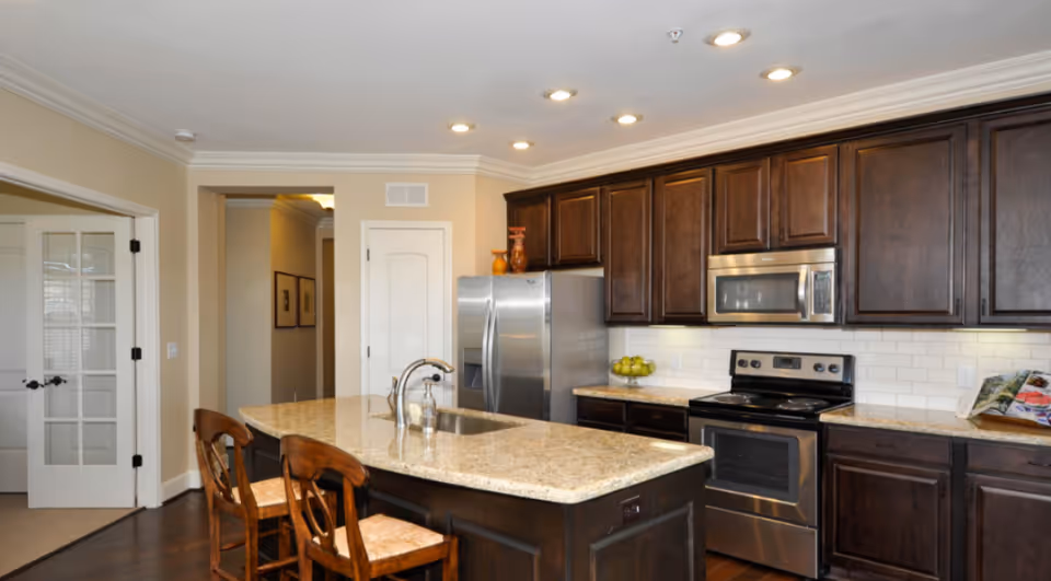Modern kitchen with dark wood cabinets, a granite island with sink and two wooden stools, and stainless steel appliances under recessed lighting.
