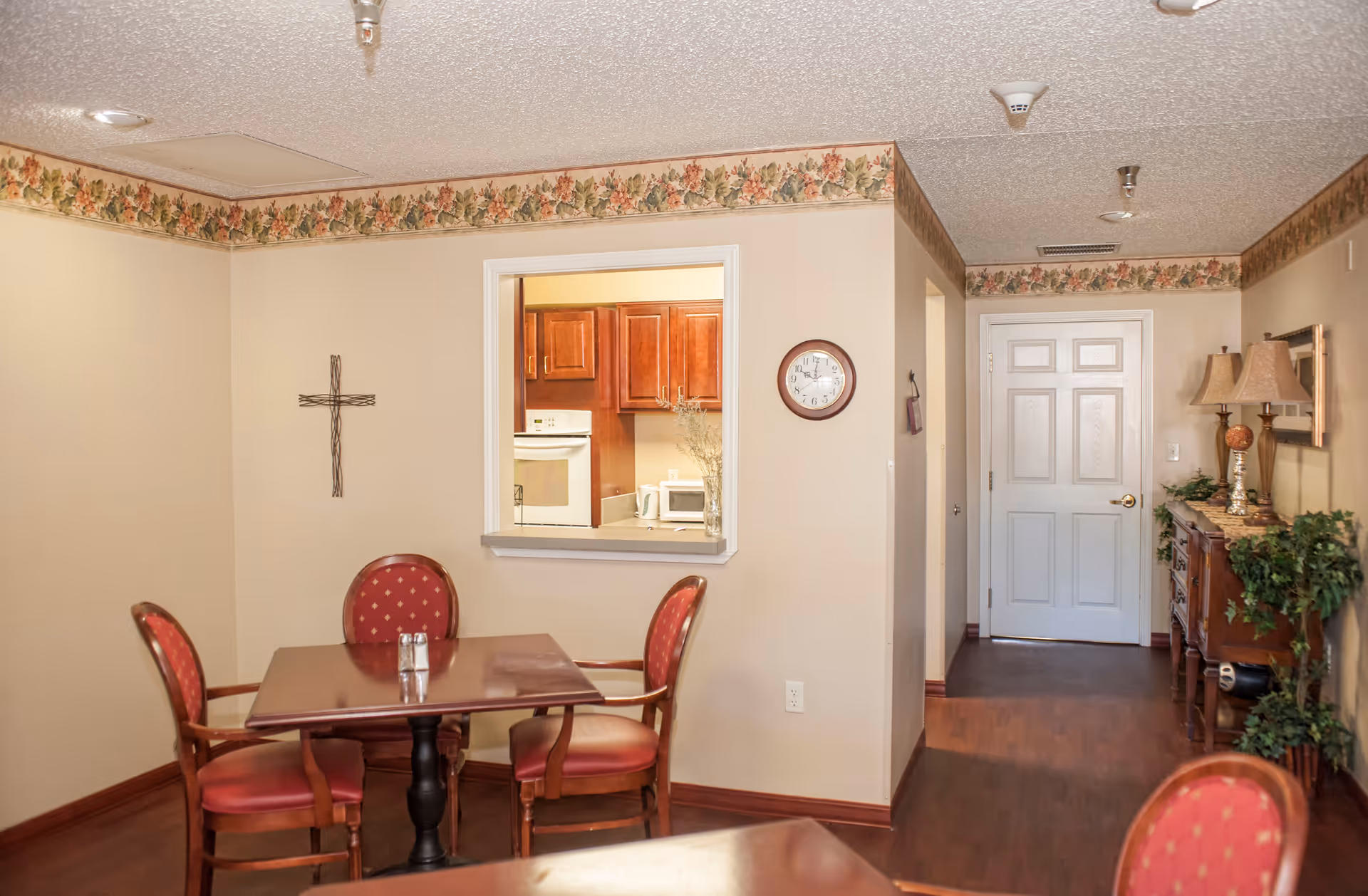 Interior view of a dining area in an assisted living facility with a square wooden table and four red cushioned chairs. A small window opening reveals a kitchen with wooden cabinets and appliances. The walls are beige with floral wallpaper border near the ceiling. A cross hangs on one wall and a clock is mounted near the kitchen window. A hallway with a white door and a wooden sideboard with lamps and plants is visible.
