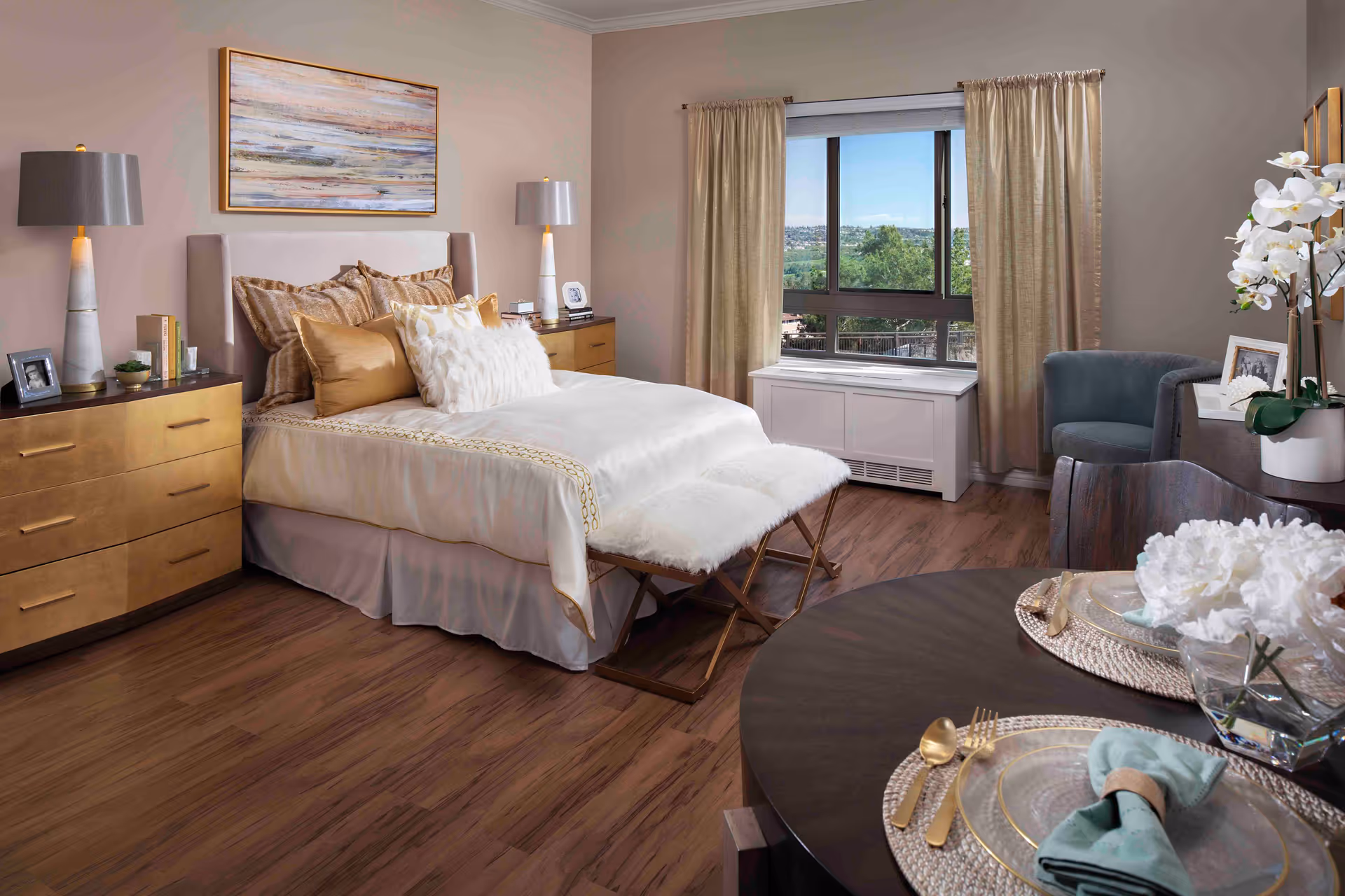 A cozy senior living bedroom with a neatly made bed featuring multiple pillows and a white fur throw. The room has wooden flooring, two matching bedside tables with lamps, a large window with beige curtains, a small seating area with a gray chair, and a round dining table set with plates, gold utensils, and white flowers.
