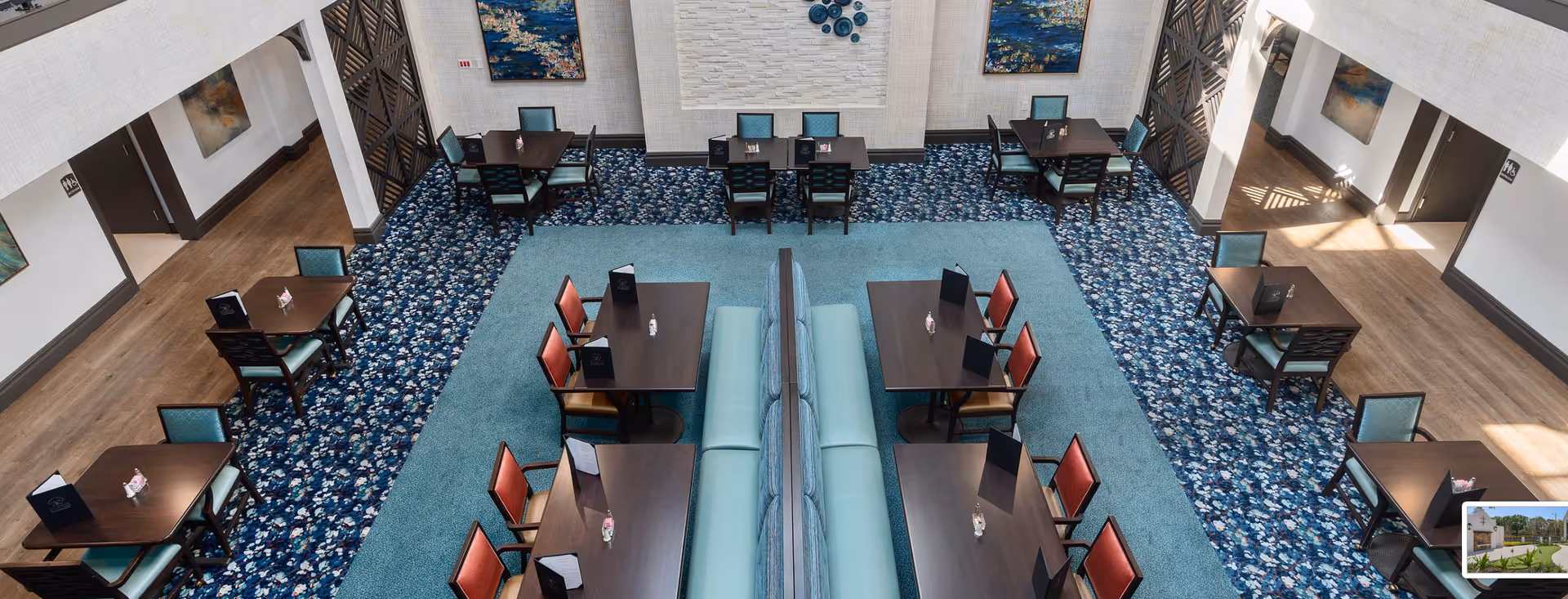 A spacious senior dining room with multiple small tables and chairs arranged on patterned carpet around a central seating area.