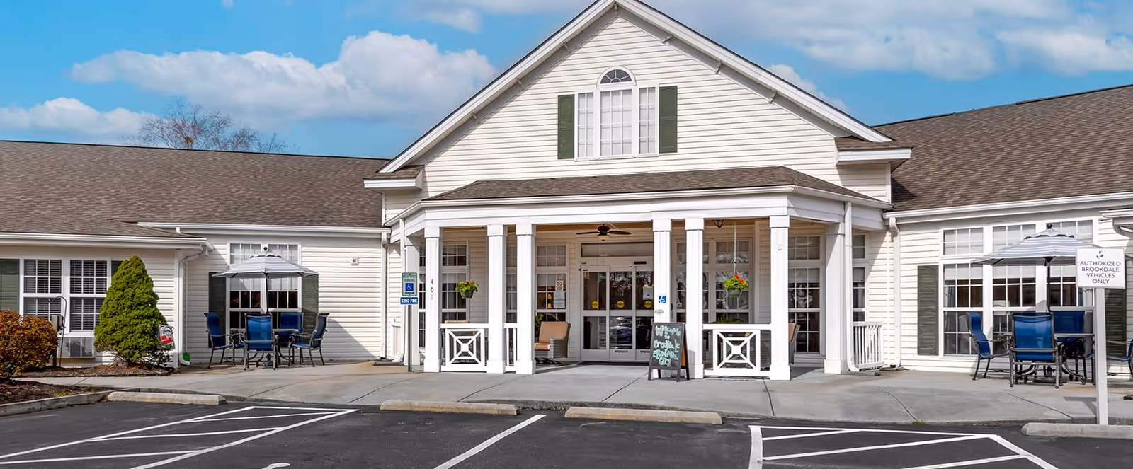 Front exterior view of Brookdale Elizabeth City facility showing a white building with green shutters, a covered entrance with columns, outdoor seating areas with tables and chairs, and a parking lot with marked spaces in front.