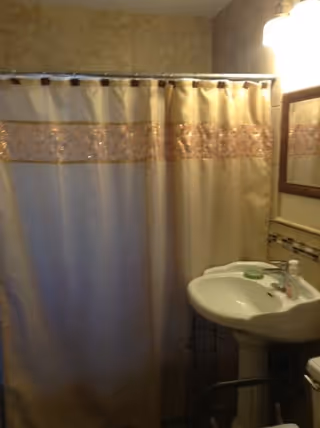 A bathroom with a beige shower curtain featuring a decorative horizontal stripe, a white pedestal sink with a green soap dish and a bottle of liquid soap, a wall-mounted mirror with a wooden frame, and a light fixture above the mirror.