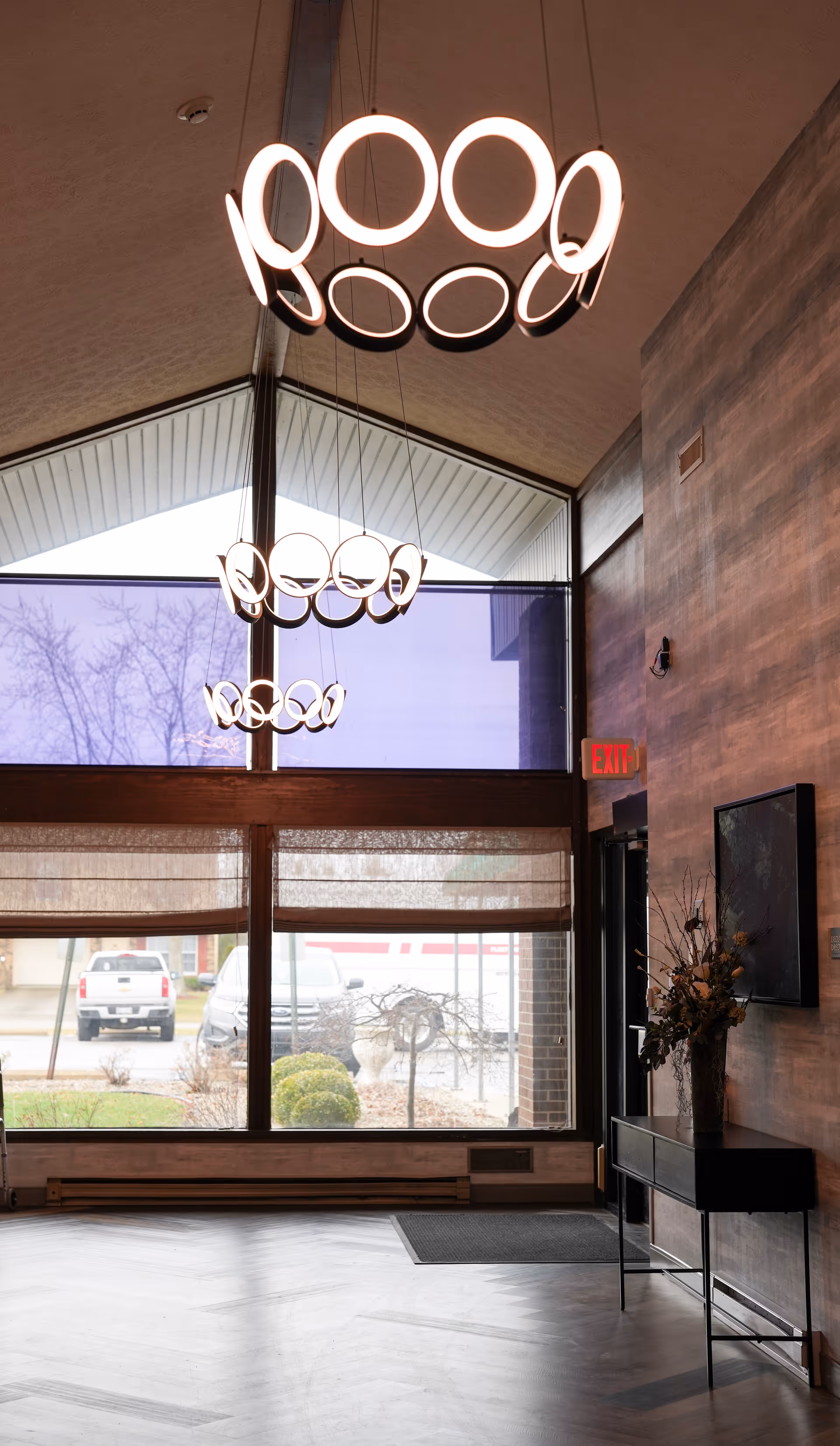 Interior view of a facility lobby with large windows showing an outdoor parking area. The ceiling is vaulted with modern circular pendant lights hanging down. There is a console table with a floral arrangement on the right side near the entrance door with an exit sign above it.