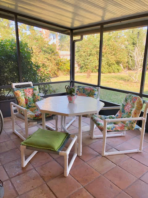 A screened-in patio area with a white hexagonal table surrounded by four white chairs with floral cushions. There is a small potted plant on the table. The patio overlooks a green garden with trees and bushes.