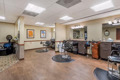 Interior view of a salon area in a senior living facility with multiple styling chairs, mirrors, and hairdressing equipment. The room has wood flooring, beige walls with framed artwork, and bright overhead lighting.
