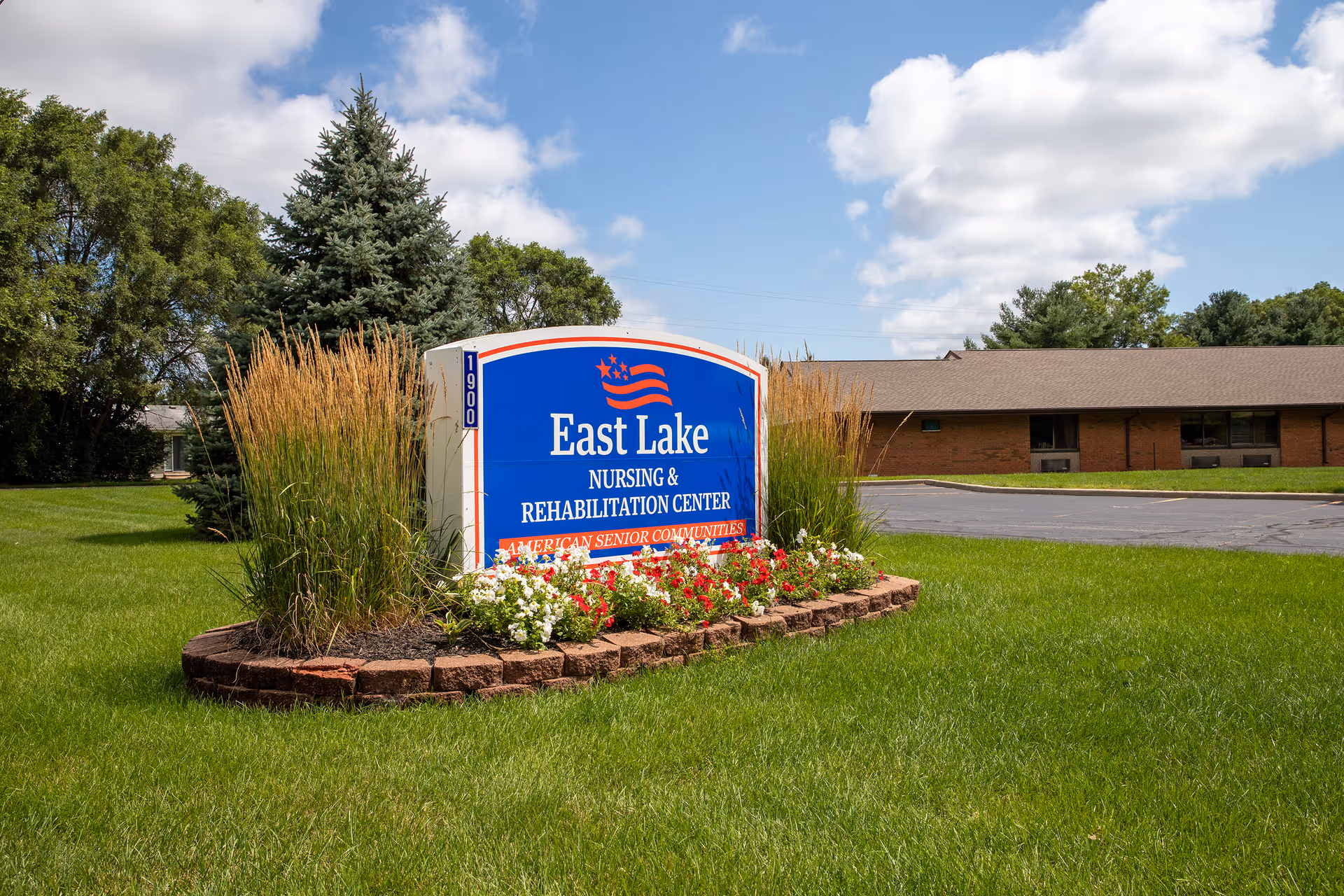 Outdoor view of the East Lake Nursing and Rehabilitation Center sign surrounded by green grass, flowers, and tall ornamental grasses with a building and trees in the background under a partly cloudy sky.
