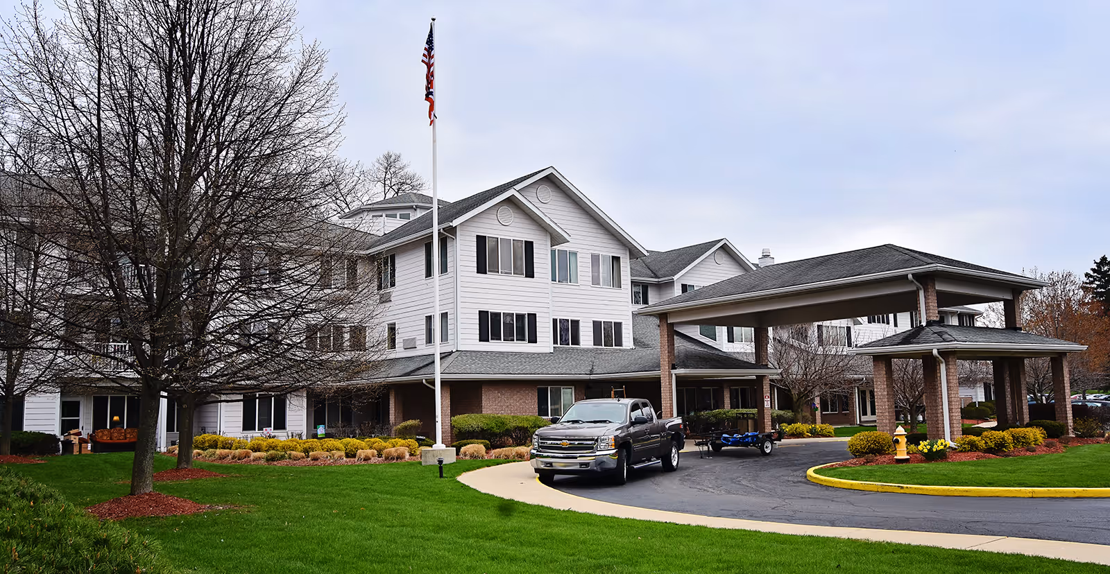 Front entrance of a white multi-story senior living building with a covered porte-cochere, flagpole, truck in the driveway, and landscaped lawn.