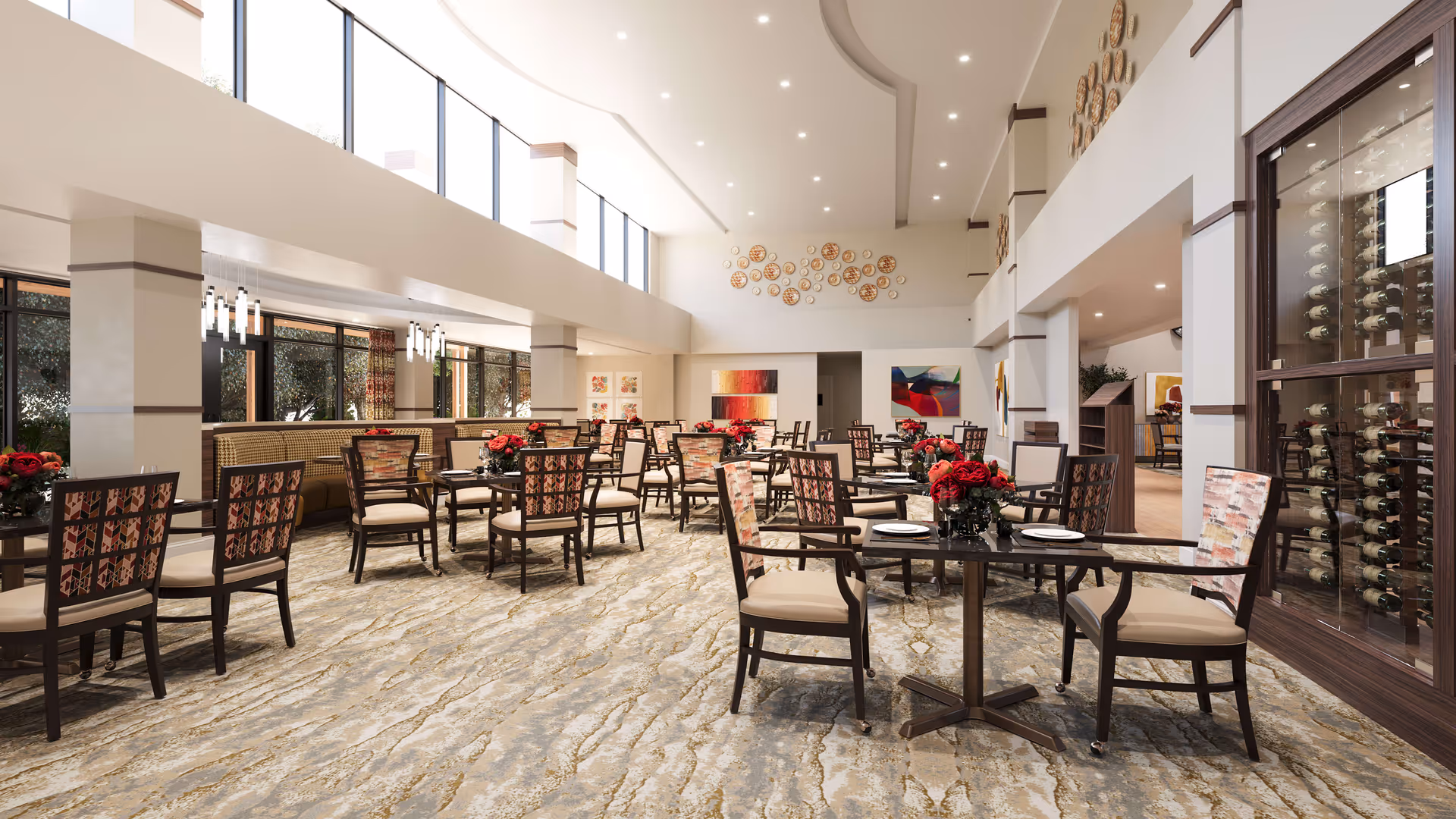 A spacious, well-lit dining room with multiple tables and chairs arranged neatly. The room features large windows near the ceiling allowing natural light to flood in. The tables are set with plates and decorated with red floral centerpieces. The walls are adorned with colorful abstract art and decorative circular wall hangings. A glass-enclosed wine rack is visible on the right side of the room.