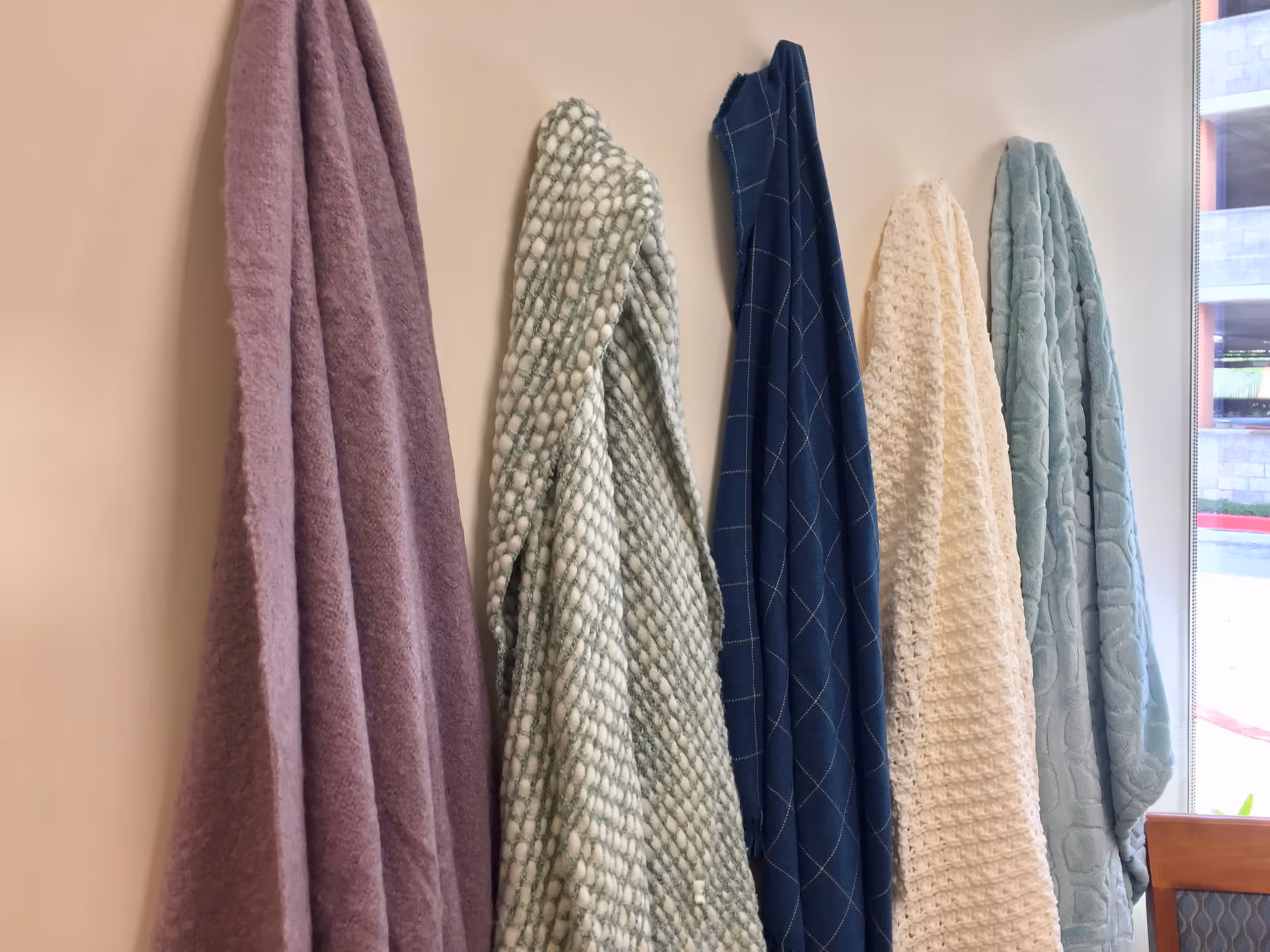 Five different textured and colored towels hanging on hooks against a beige wall next to a window with a partial view of the outside and a wooden chair with patterned upholstery.