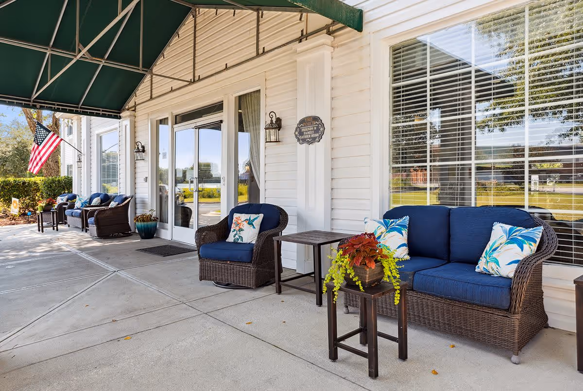 Outdoor seating area at the entrance of Florence Place featuring wicker chairs and sofas with blue cushions and decorative pillows, small tables with potted plants, an American flag, and large windows with blinds under a green awning.