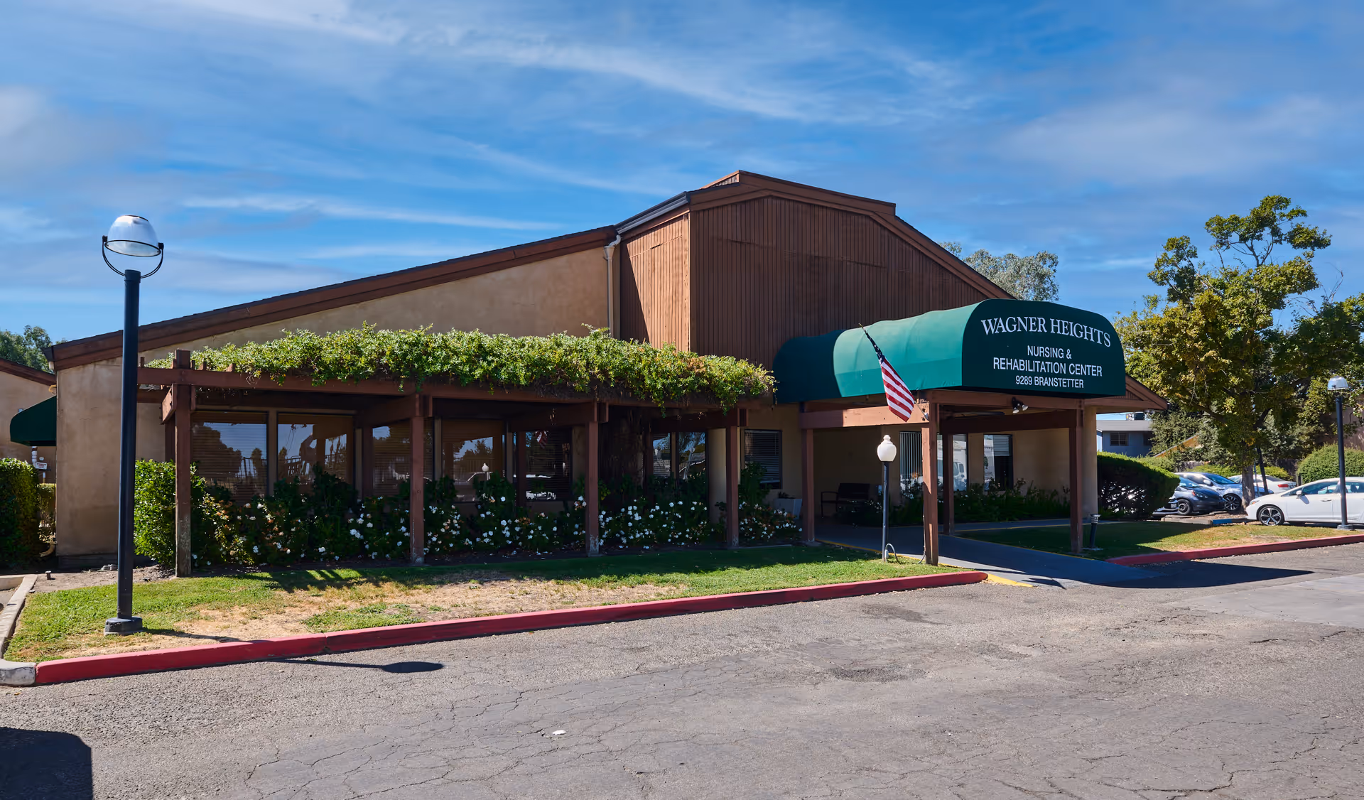 Exterior view of Wagner Heights Nursing & Rehabilitation Center building with a green awning over the entrance, an American flag, a pergola with greenery and flowers, a lamp post, and parked cars in the background under a blue sky.