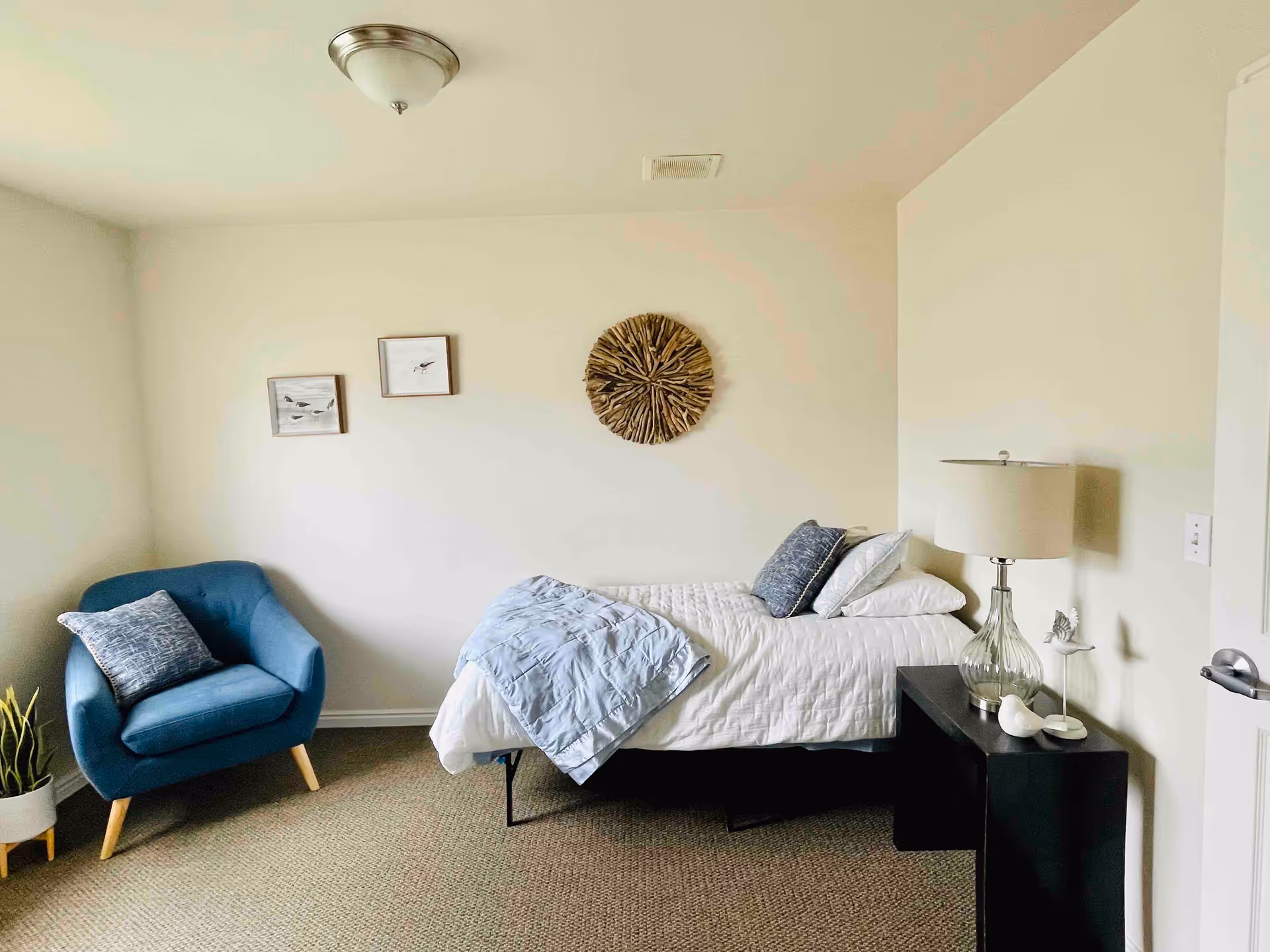 A simple, clean bedroom with a single bed made with white bedding and a light blue blanket. There are two blue pillows on the bed. To the right of the bed is a black nightstand with a glass lamp and decorative bird figurines. On the left side of the room is a blue armchair with a patterned cushion and a potted plant on the floor beside it. The walls are light-colored with two small framed pictures and a round wooden wall decoration above the bed.