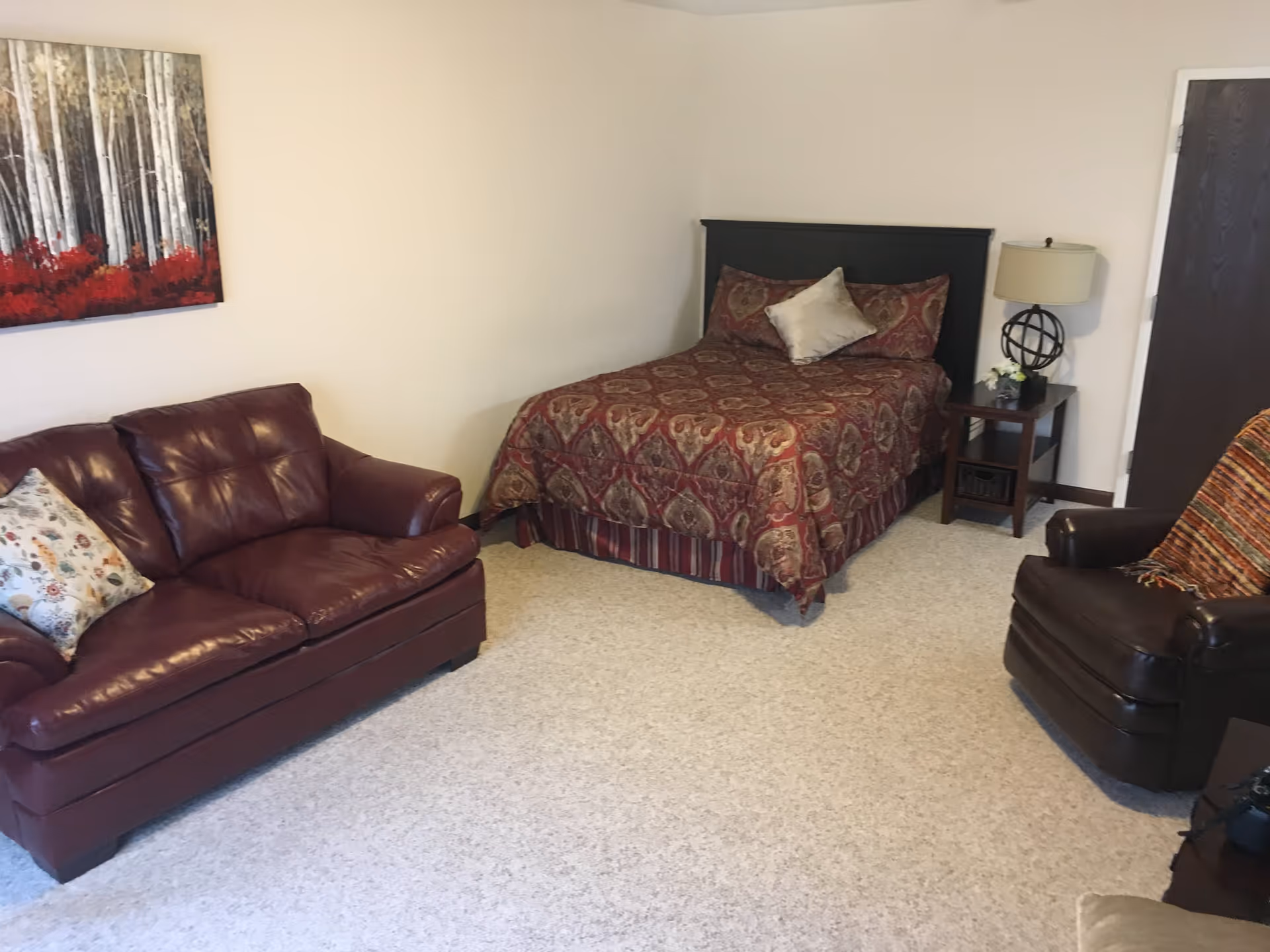 A cozy senior living room with a patterned bed, a dark leather sofa with a floral pillow, a dark leather armchair with a colorful throw, a wooden side table with a lamp and small plant, and a painting of birch trees with red foliage on the wall.