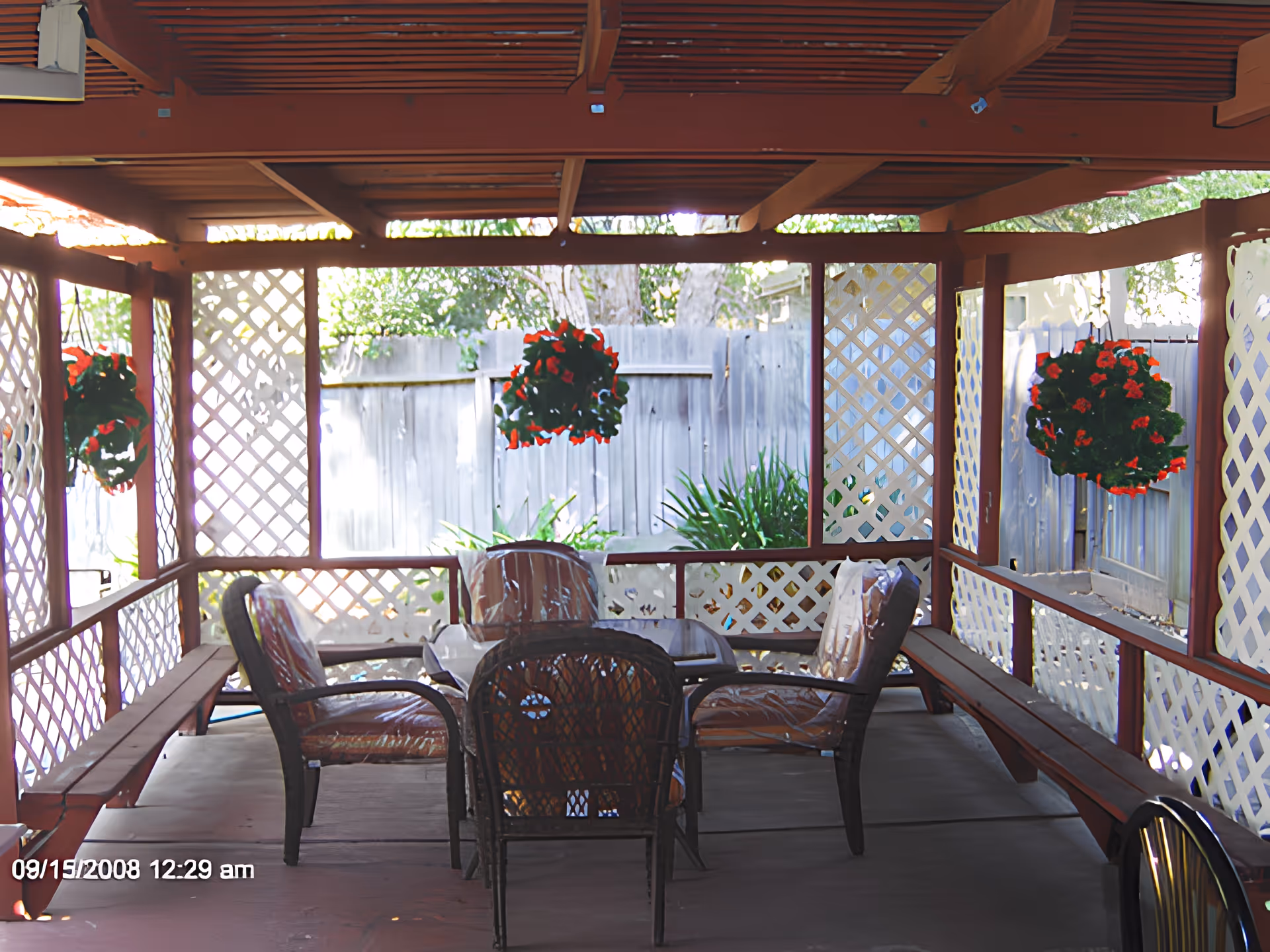 Covered outdoor patio with a table, chairs, lattice walls and hanging red flower baskets.