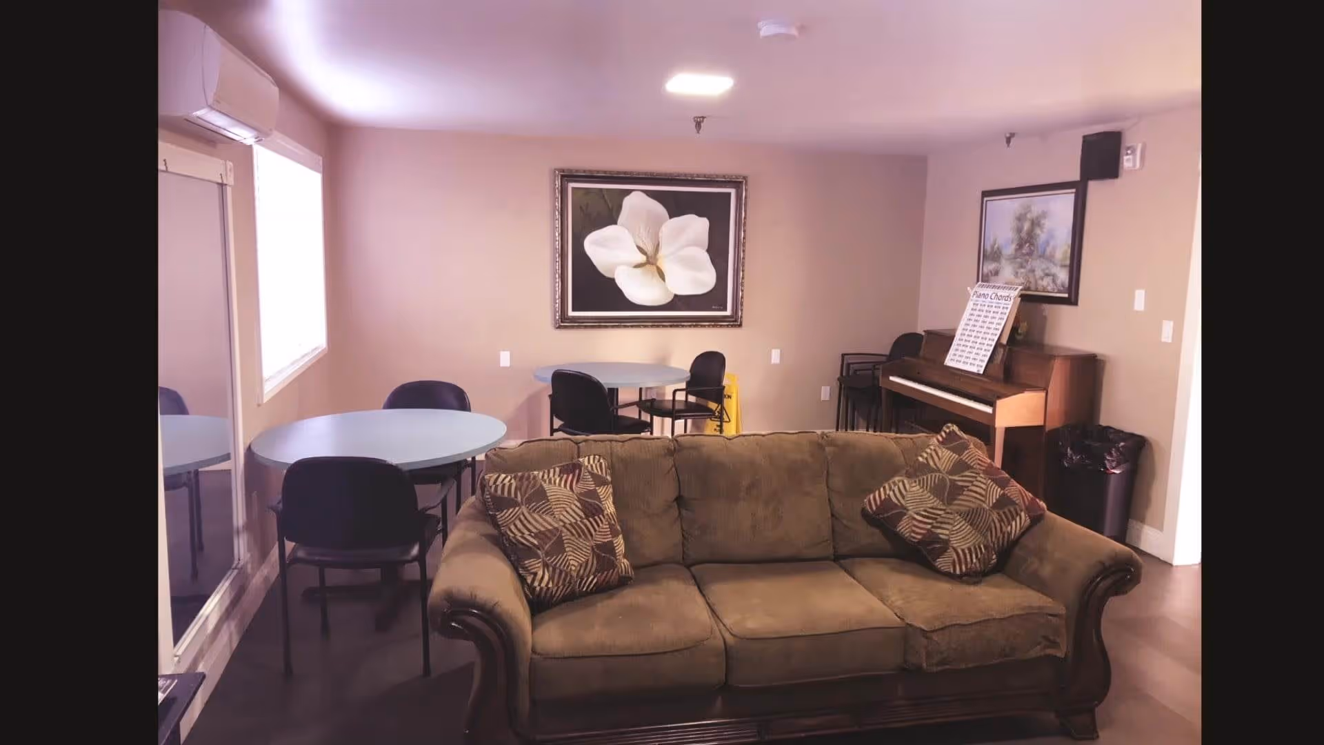 A cozy living room area with a brown sofa adorned with two patterned cushions. Behind the sofa, there are two round tables each surrounded by black chairs. On the right side, there is an upright piano with a sheet of piano chords on the music stand and a framed painting above it. The walls are beige, and there is a large framed picture of a white flower on the wall. A window with blinds is on the left side, and a trash bin is visible near the piano.