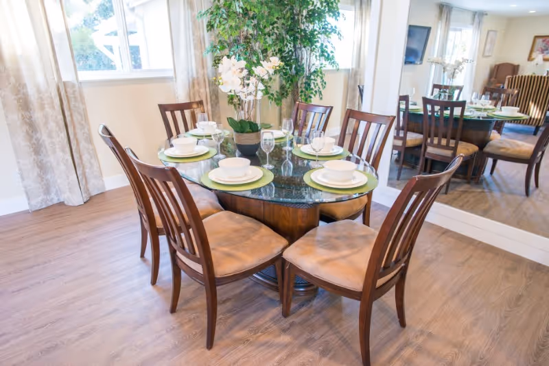 A round glass dining table set for six with white bowls, plates, and glasses on green placemats. The table is surrounded by six wooden chairs with cushioned seats. A potted orchid plant is in the center of the table. The room has wooden flooring, a large window with curtains, and a large mirror reflecting the dining area.