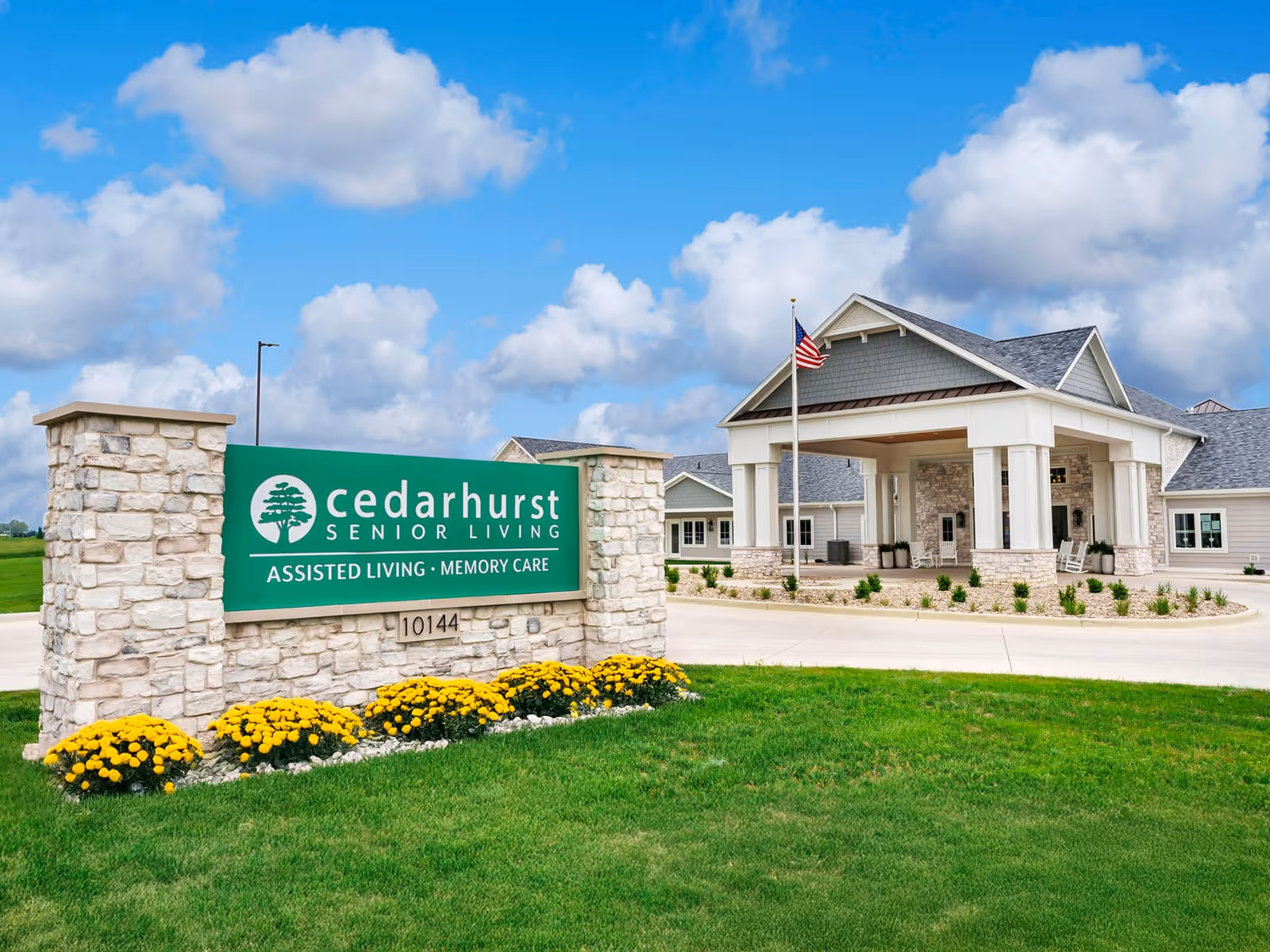 The front entrance and sign for Cedarhurst Senior Living with landscaped lawn, yellow flowers, driveway, and an American flag.