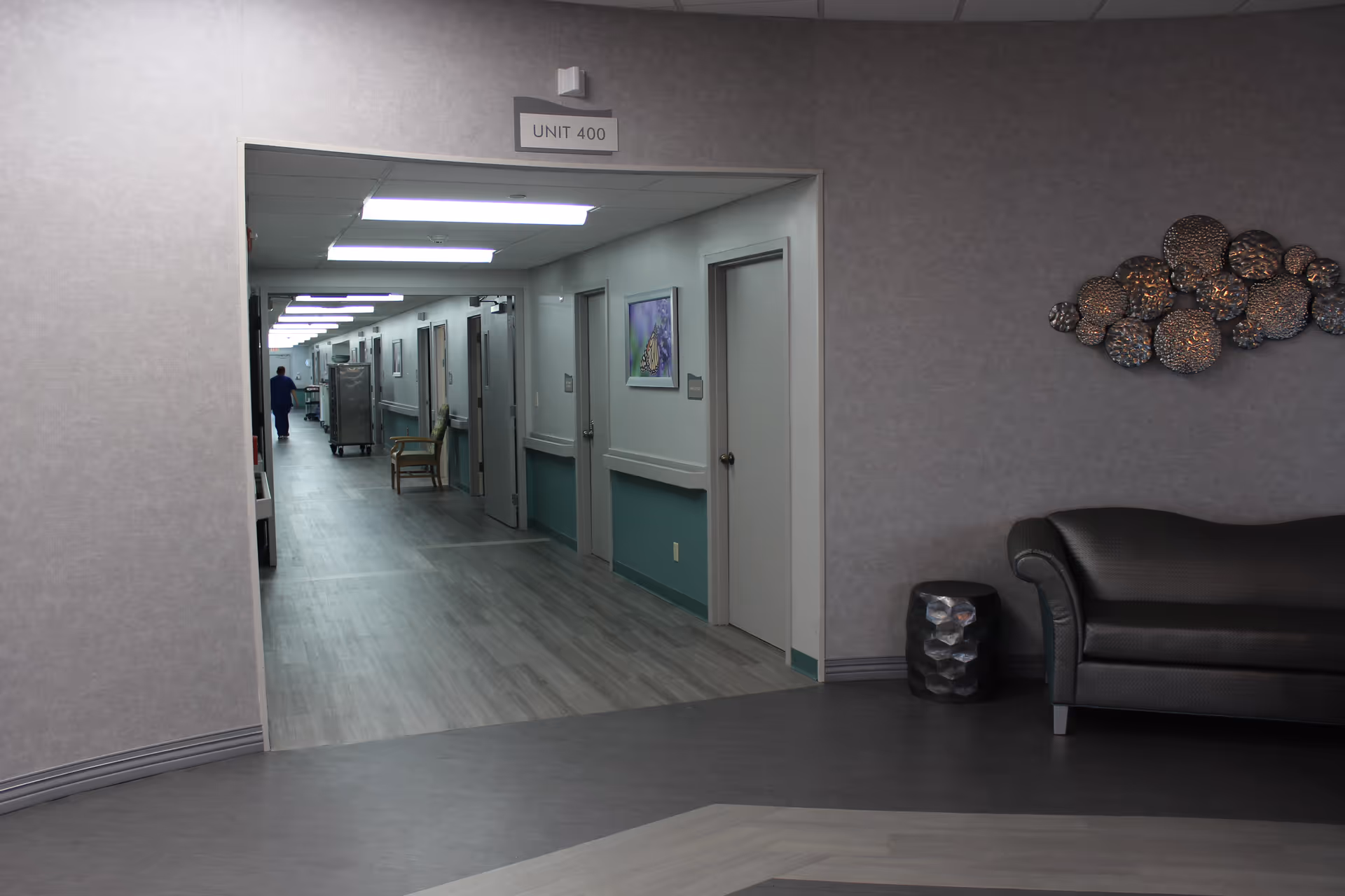 A hallway inside a senior living facility labeled Unit 400. The hallway has several doors on the right side, a chair, and a person in blue scrubs walking away in the distance. The foreground shows a seating area with a dark couch and a decorative wall piece made of circular metallic elements.