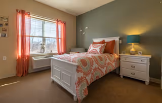 A cozy bedroom with a white wooden bed frame, a colorful patterned bedspread in shades of orange and teal, and matching pillows. There is a white nightstand with a lamp and a small picture frame beside the bed. The room has beige walls, a large window with orange curtains, and a beige carpeted floor.