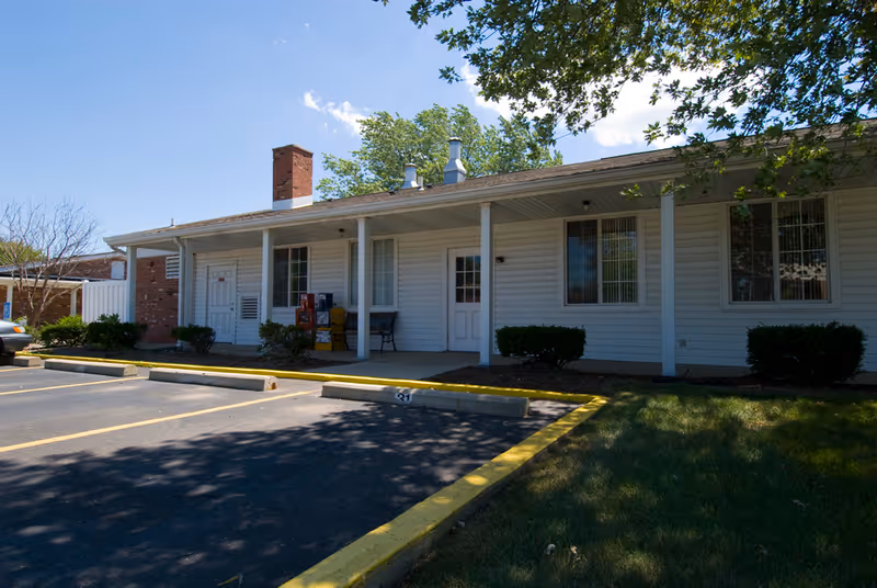 Exterior view of a single-story building with white siding and a covered porch supported by white columns. There are several windows and doors along the front, with a brick chimney visible on the left side. The building is surrounded by a parking lot with marked spaces and some greenery, including bushes and a tree casting shadows on the ground.