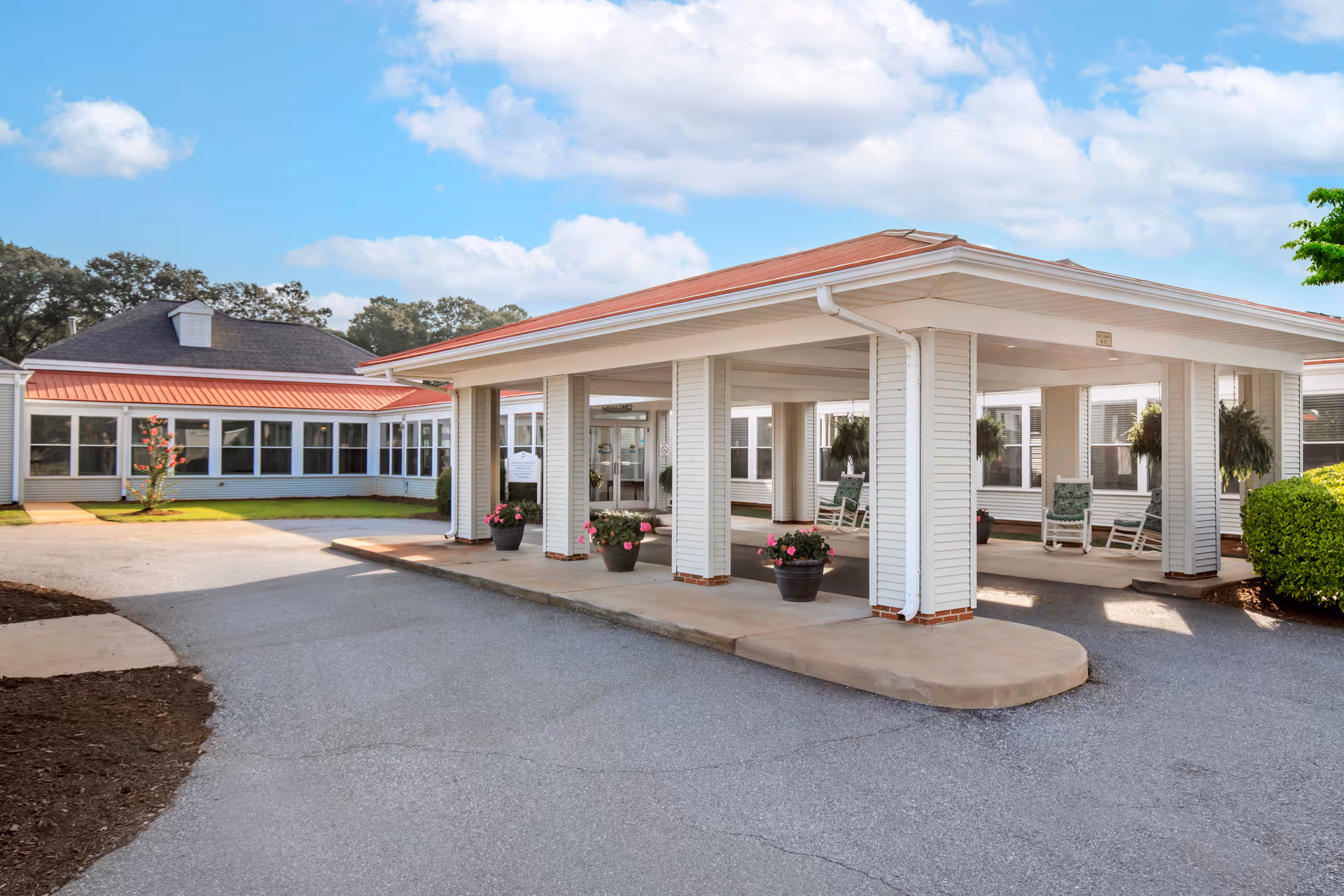 Covered entrance and porte-cochere of a senior living facility with potted flowers, rocking chairs, and a driveway.