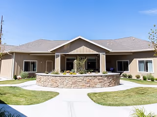 Exterior view of a single-story building with beige siding and a gray roof, featuring a circular stone planter with greenery in front, surrounded by well-maintained grass and concrete walkways under a clear blue sky.