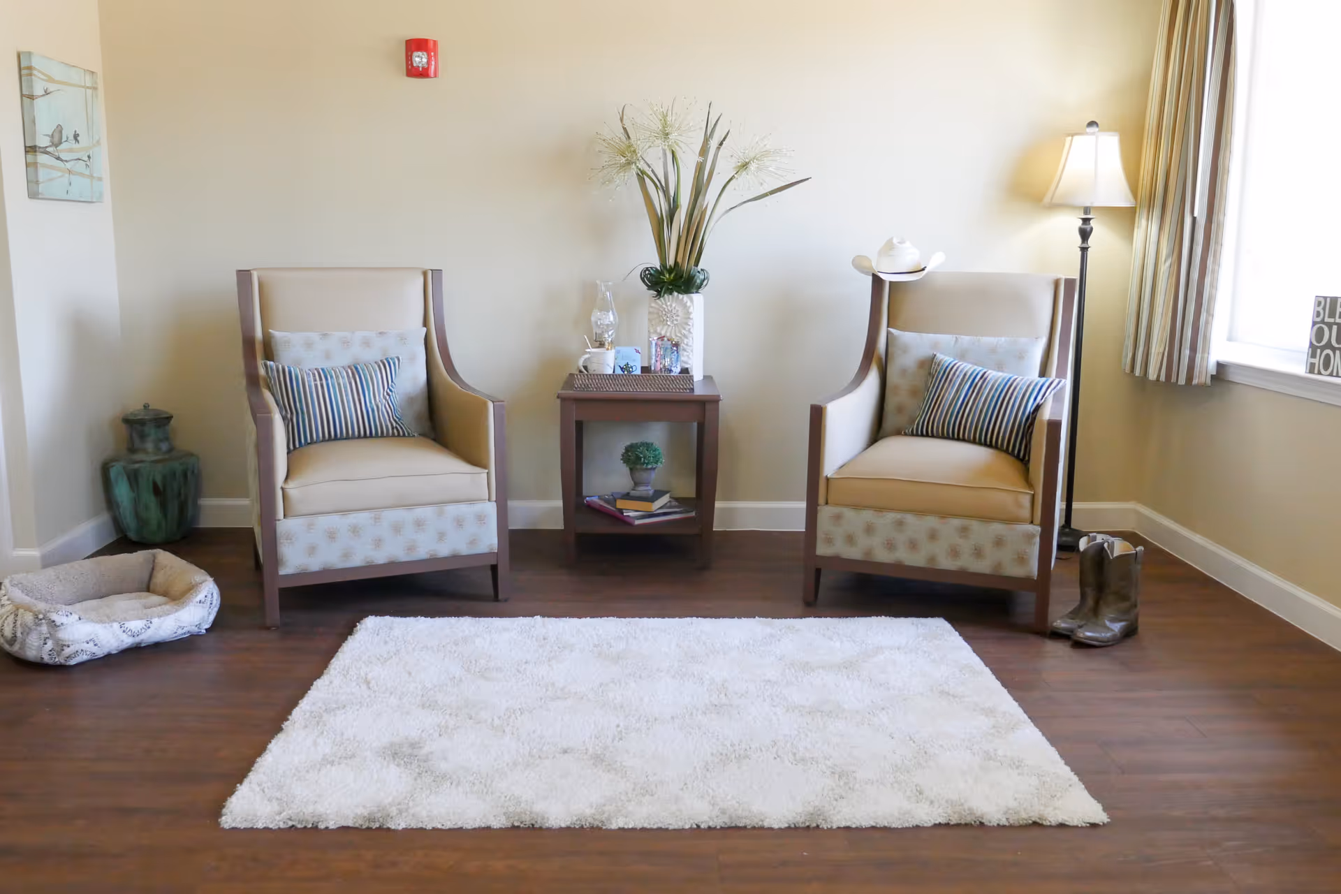 A cozy sitting area with two beige armchairs featuring patterned cushions, a small wooden side table with decorative items and a vase of flowers, a floor lamp, a white rug on wooden flooring, a pet bed on the left, and a pair of cowboy boots next to the right chair near a window with striped curtains.