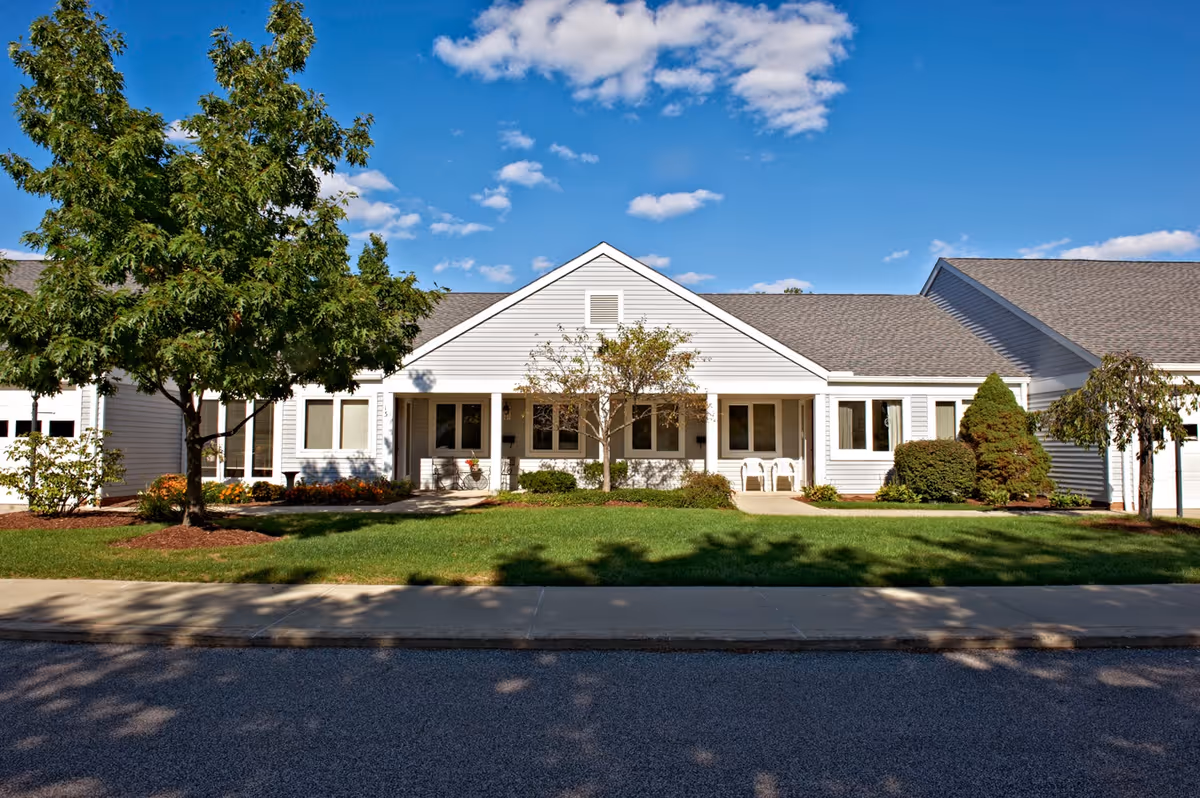 Front exterior of a single-story white retirement campus building with a lawn, trees, and a walkway.