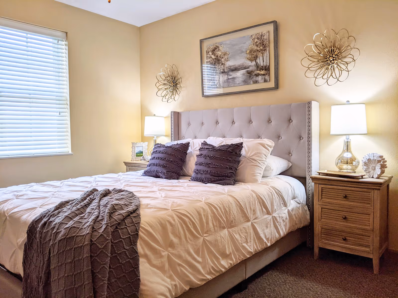 A cozy bedroom with a large bed featuring a tufted headboard, white bedding, and two purple decorative pillows. There is a gray textured throw blanket draped over the foot of the bed. On each side of the bed are wooden nightstands with lamps, decorative items, and a framed picture. The walls are painted beige and decorated with a framed landscape painting and two metal wall sculptures. A window with closed blinds is on the left side of the room.
