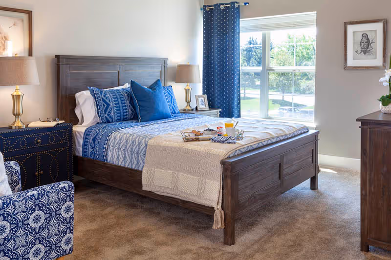 Sunlit bedroom with a wooden bed dressed in blue patterned bedding, matching blue curtains, nightstands with lamps, and a window overlooking greenery.