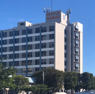 Multi-story Hospital Damas building with rooftop antennas, trees in the foreground, and a clear blue sky.