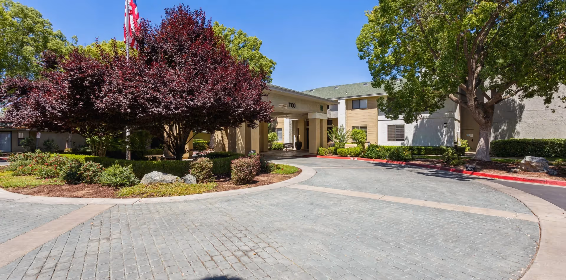 Entrance to a senior living facility with a circular driveway, landscaped garden with bushes and trees including a large tree with dark red leaves, and an American flag on a flagpole. The building is light-colored with a covered entryway and windows.