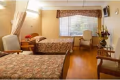 A cozy bedroom in a senior living facility with two single beds covered in floral bedspreads, a window with blinds and a valance, two armchairs, a small table with a flower vase, and a wall-mounted television.