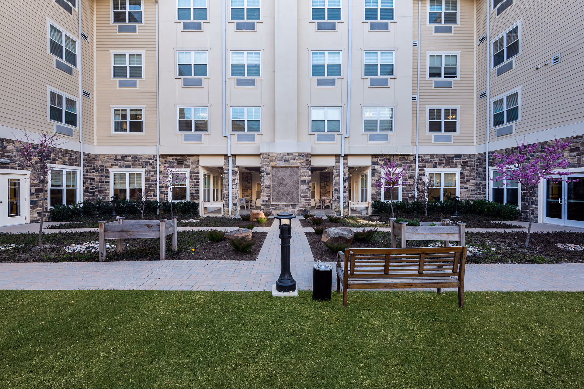 Courtyard with benches, a paved walkway, planted beds and a multi-story building facade with many windows.